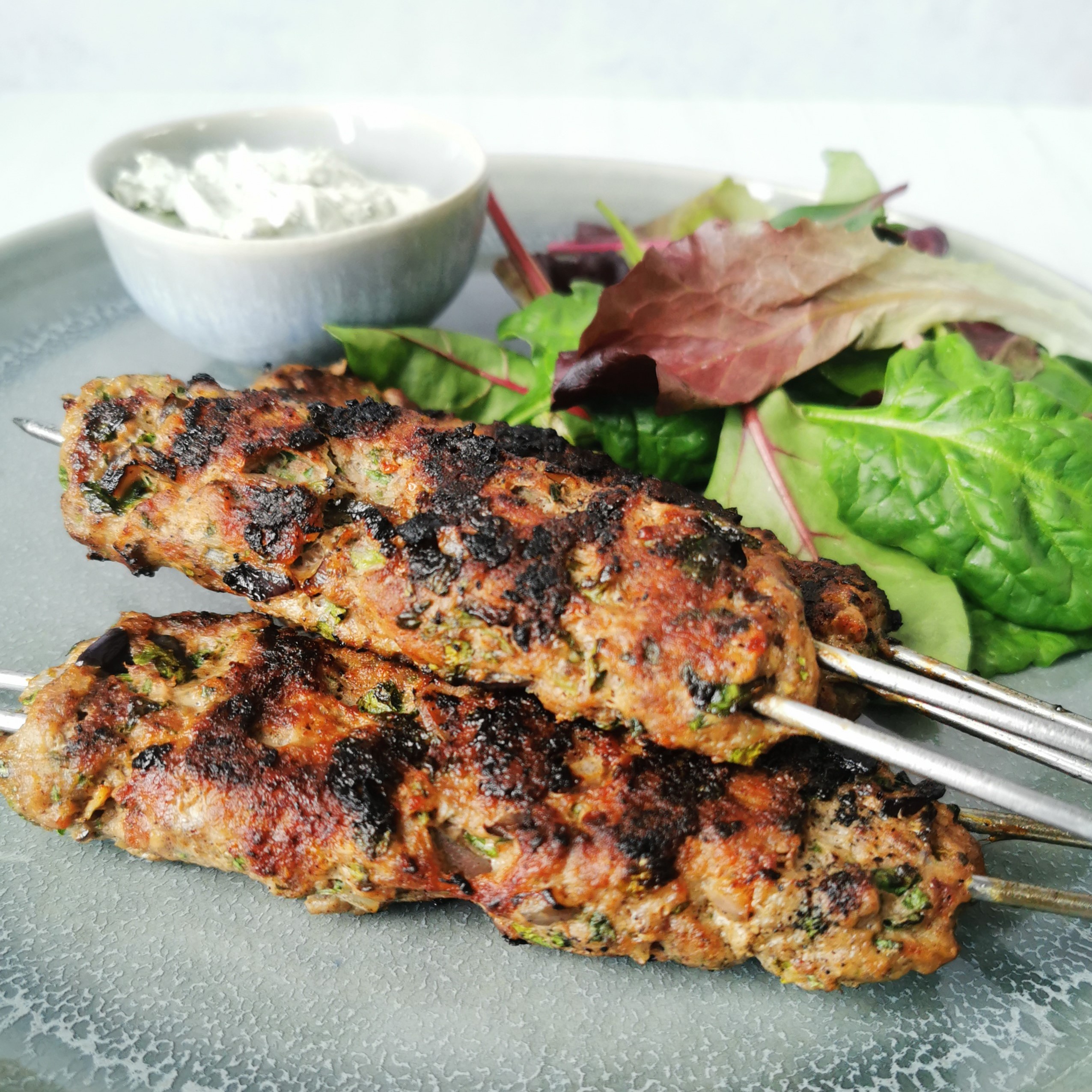 Lamb koftas on metal skewers with sald and a small bowl of mint yoghurt on a grey plate