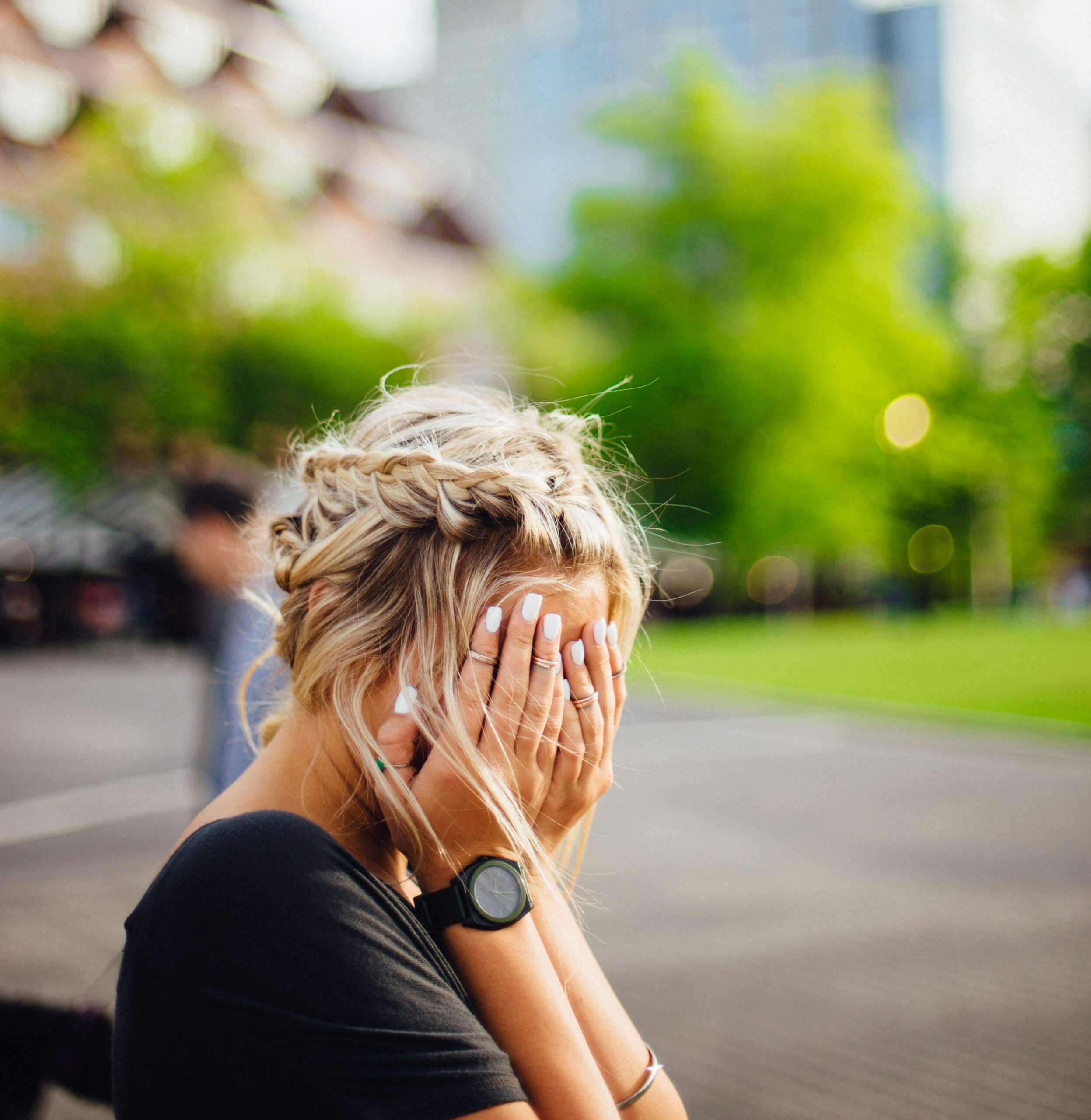 a blonde woman wit her hands covering her face in despair, sitting outside.