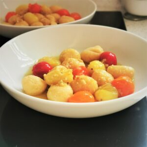 Two white bowls of gnocchi, one in front of the other. Balls of gnocchi with red, orange and yellow baby plum tomatoes with garlic butter and oil. The background is black