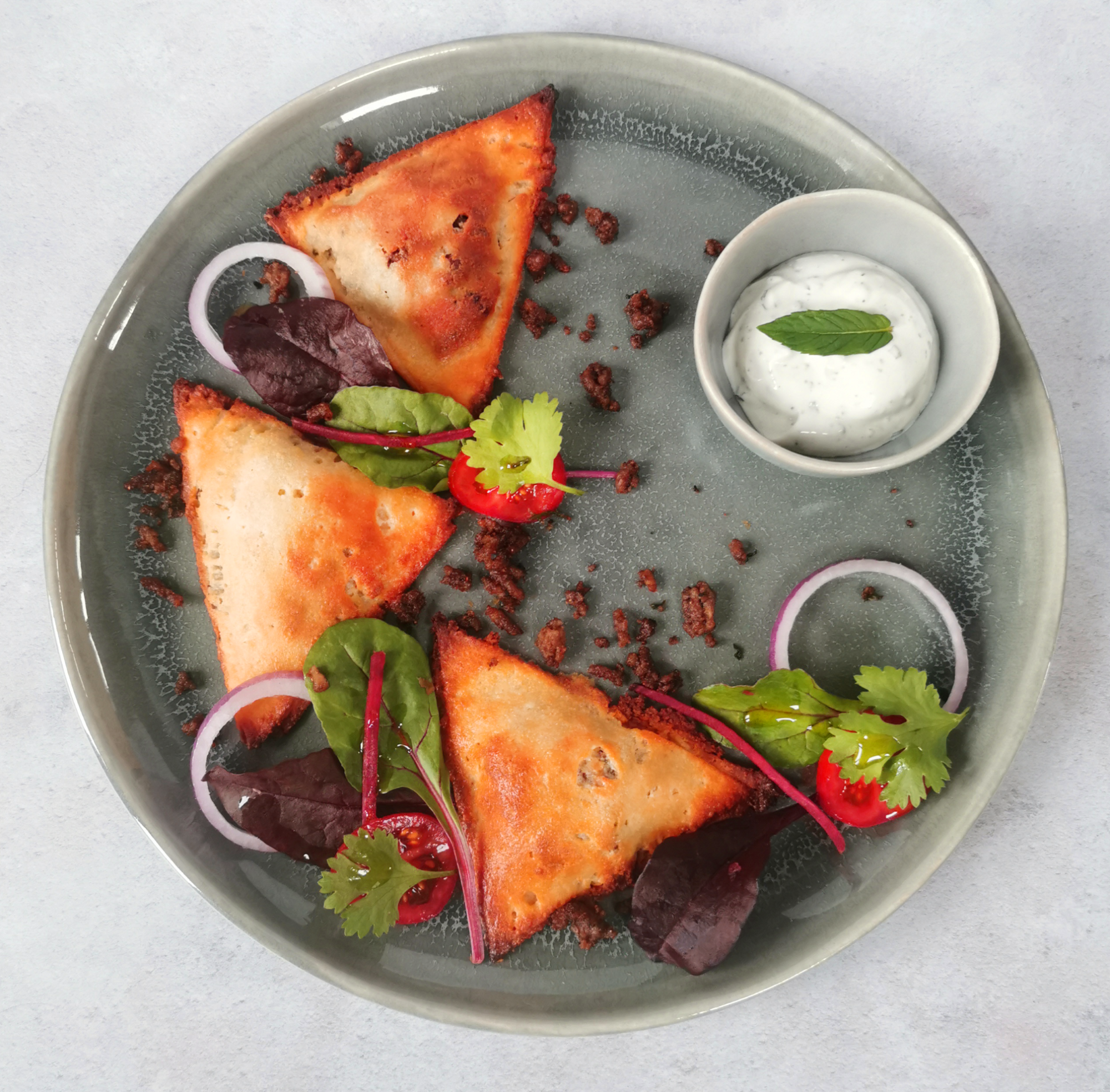 grey plate on a white background with 3 lamb samosas, a bowl of mint yoghurt and salad garnish