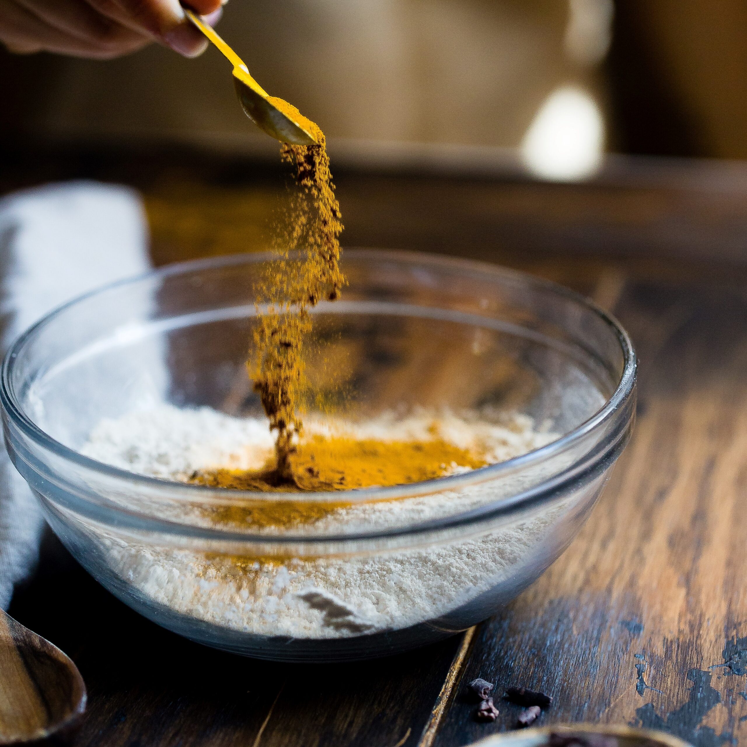 a hand with spoon sprinkling turmeric into a glass bowl of ground almonds sitting on a wooden surface