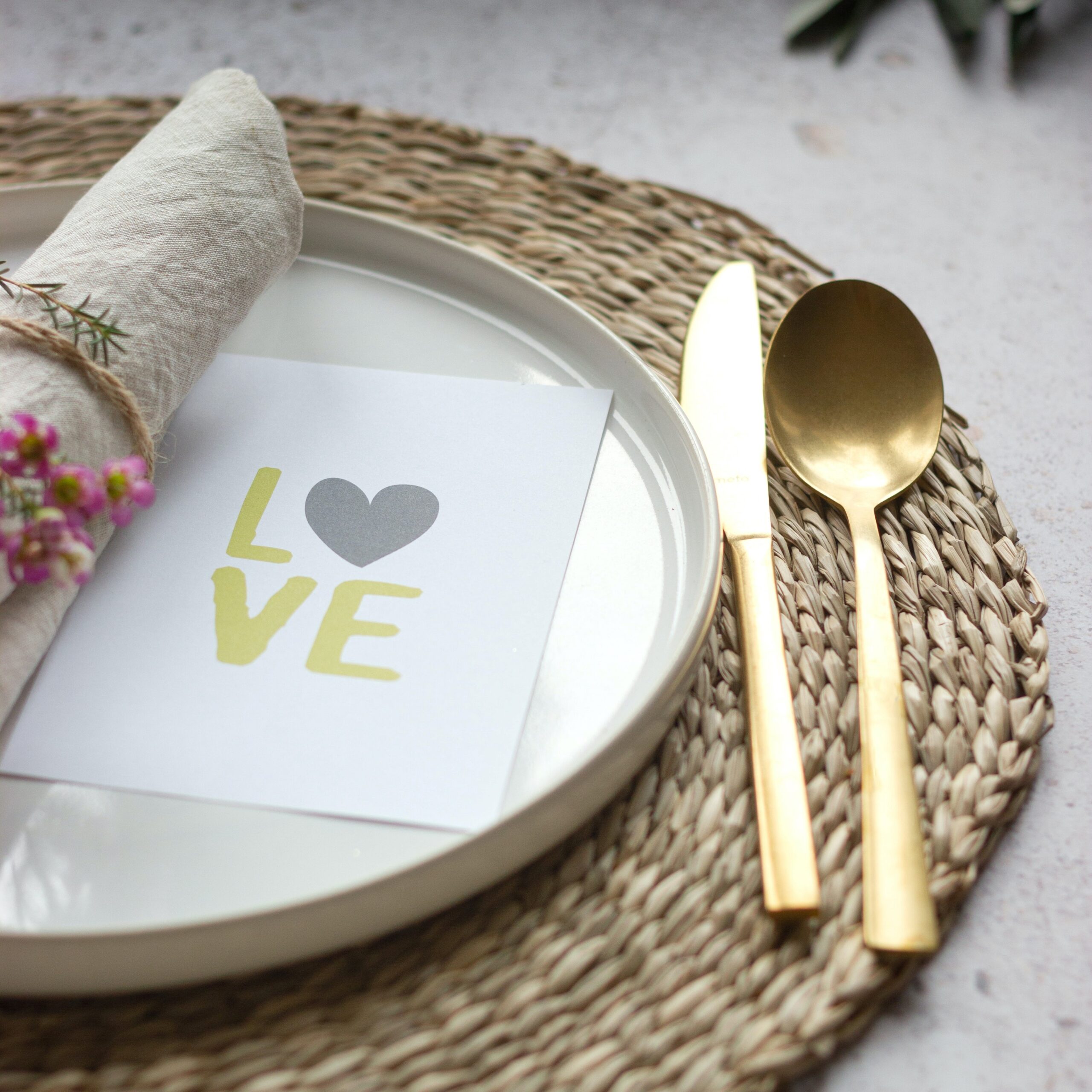 A rafia placemat with a white plate upon which is a card saying love, a napkin with flowers as a napkin ring and a gold knife and spoon.