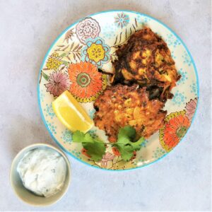 floral plate on a mottled background with two onion bhaji, a lemon wedge, a sprig of coriander and a small bowl of mint yoghurt