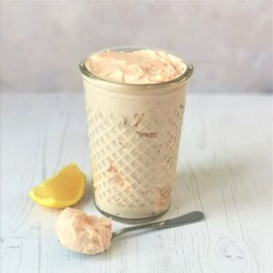 a glass jar filled with pale pink taramasalata with a spoon full and a wedge of lemon on a white washed wood surface