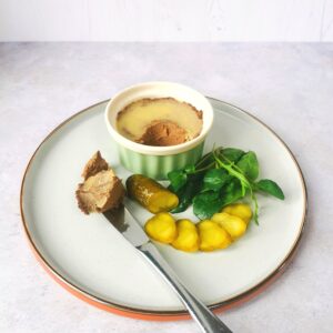 a ramekin of chicken liver pate on a plate with a knife and sliced haimisha pickles and salad