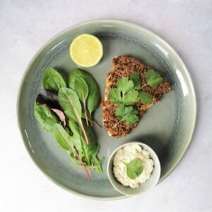 Plan view of a piece of crumbed cod on a grey plate with half a lime, salad and a small bowl of tartare sauce