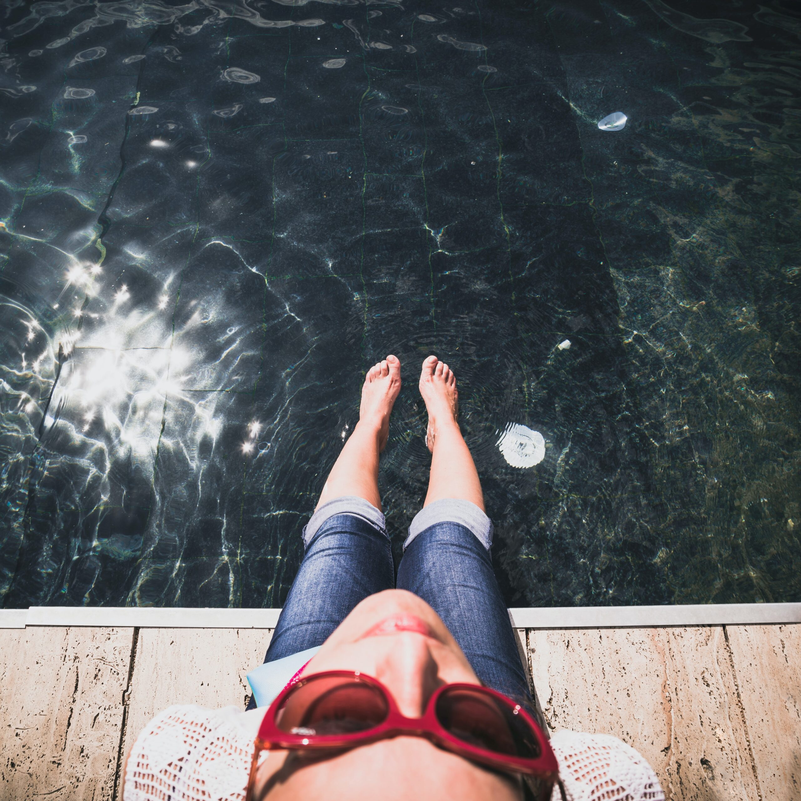 An aerial view of a lady wearing jeans rolled to her knees and red sunglasses with feet dangling in the water face up to the sun