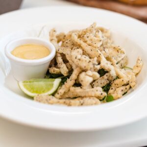 a pile of fried golden calamari on a white bowl with a small pot of mayo and a lime wedge