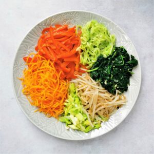 six bibimbap vegetables cooked and arranged on a grey plate on a mottle grey background