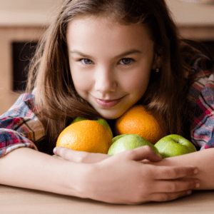 girl sitting at a table with her arms around apples and oranges