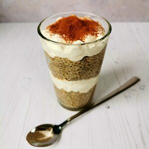 a glass with layers of coffee infused chia seeds and whipped cream, topped with cocoa powder and shown with a long sundae spoon