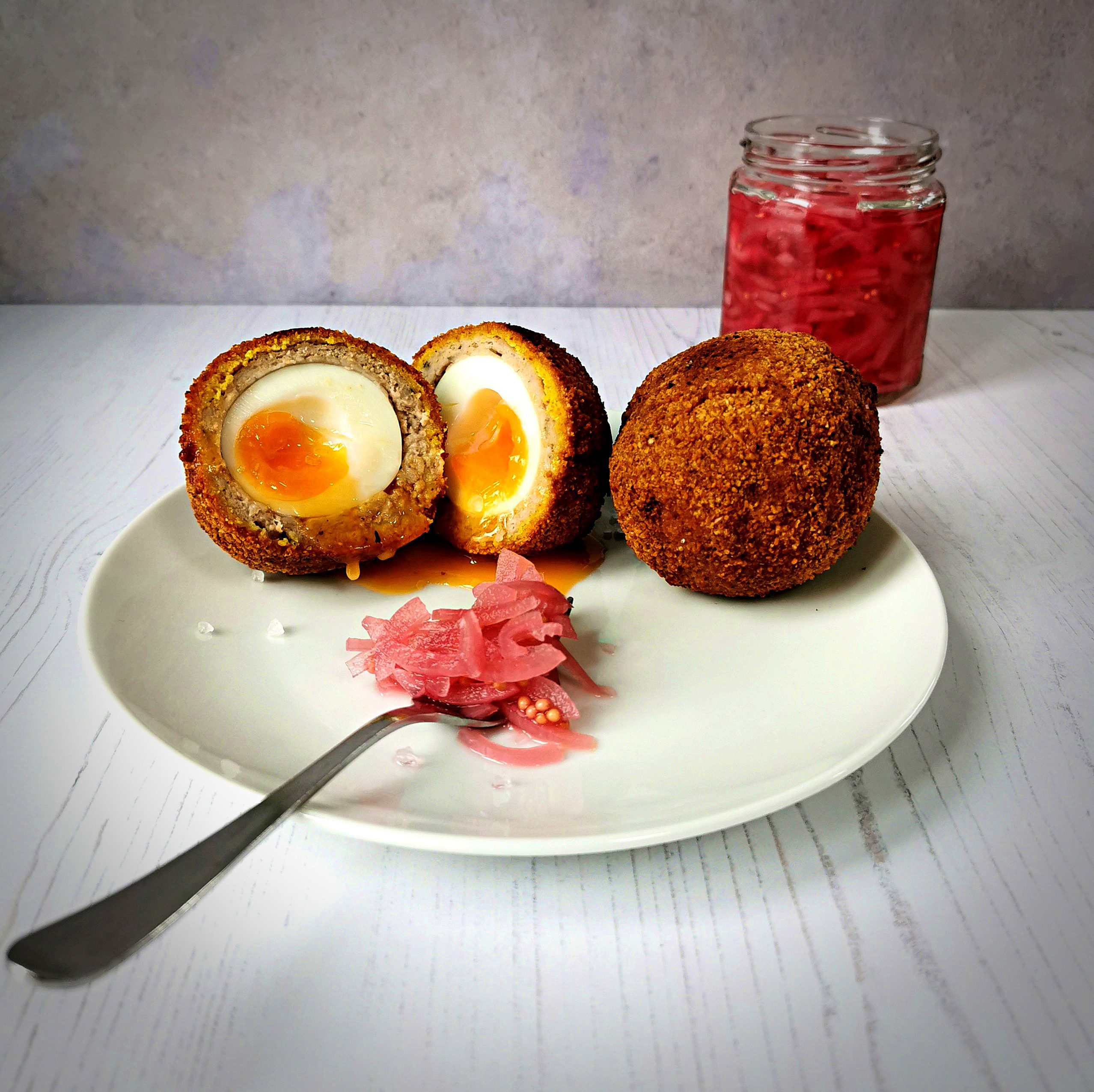 A scotch egg cut in half with a golden runny yolk. Plus a whole scotch egg on a plate with a forkful of pink pickled onions and a jar behind.
