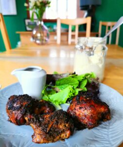 3 pieces of jerk chicken on a grey plate, with a small jug of sauce, salad and a jar of mayo behind. sitting on a wooden table