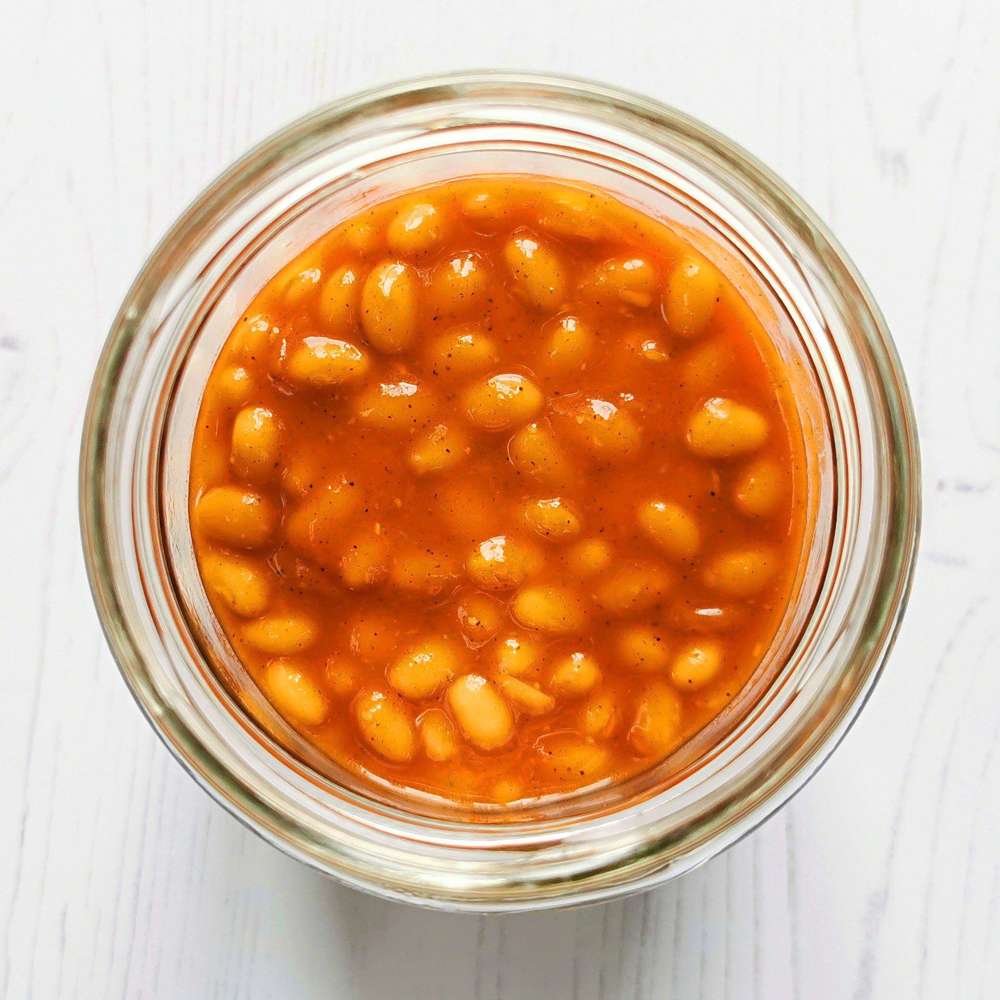 A plan view of orange baked beans in a round glass jar on a white wood background